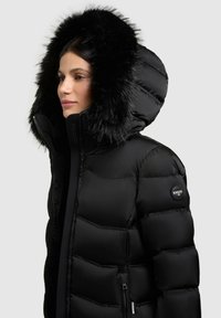 Sort pufferjakke med en faux fur hætte, quiltet tekstur, lynlås lukning og et logo patch på venstre ærme.