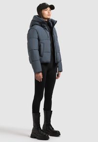 Dikke, cropped jas in gedempt blauw met een glanzende afwerking, gecombineerd met zwarte leggings en kniehoge veterschoenen. De pet voegt een casual tintje toe.