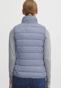 Hellblau gesteppte Weste mit hohem Kragen, horizontalem Steppmuster und ohne Ärmel, getragen über einem grauen Langarmshirt.