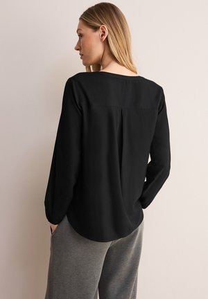Zwarte blouse met lange mouwen en een ronde halslijn, zachte textuur en een rugplooi-detail. Combinatie met grijze joggingsbroek.