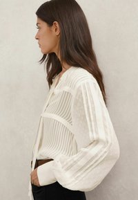 Valge, pika varrukaga pluus, millel on vertikaalsed triibud ja täpiline tekstuur, lõdva lõikega esikinnis ning pruun vööga stiilitud.