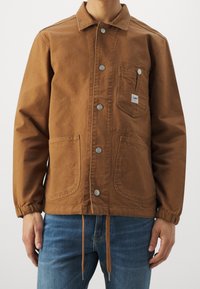 Man som bär en brun knappjacka med tre framfickor och elastiska ärmar, tillsammans med blå denimjeans.