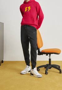 Czerwona bluza z kapturem z żółtym wzorem błyskawicy, czarne spodnie dresowe z logo Nike oraz białe sneakersy Air Jordan na żółtej podłodze.