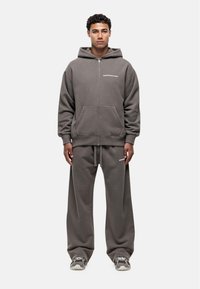 Grijze rits hoodie met voorzakken en logo. Gecombineerd met losse grijze sweatpants. Beide gemaakt van zacht materiaal. Model draagt sneakers.