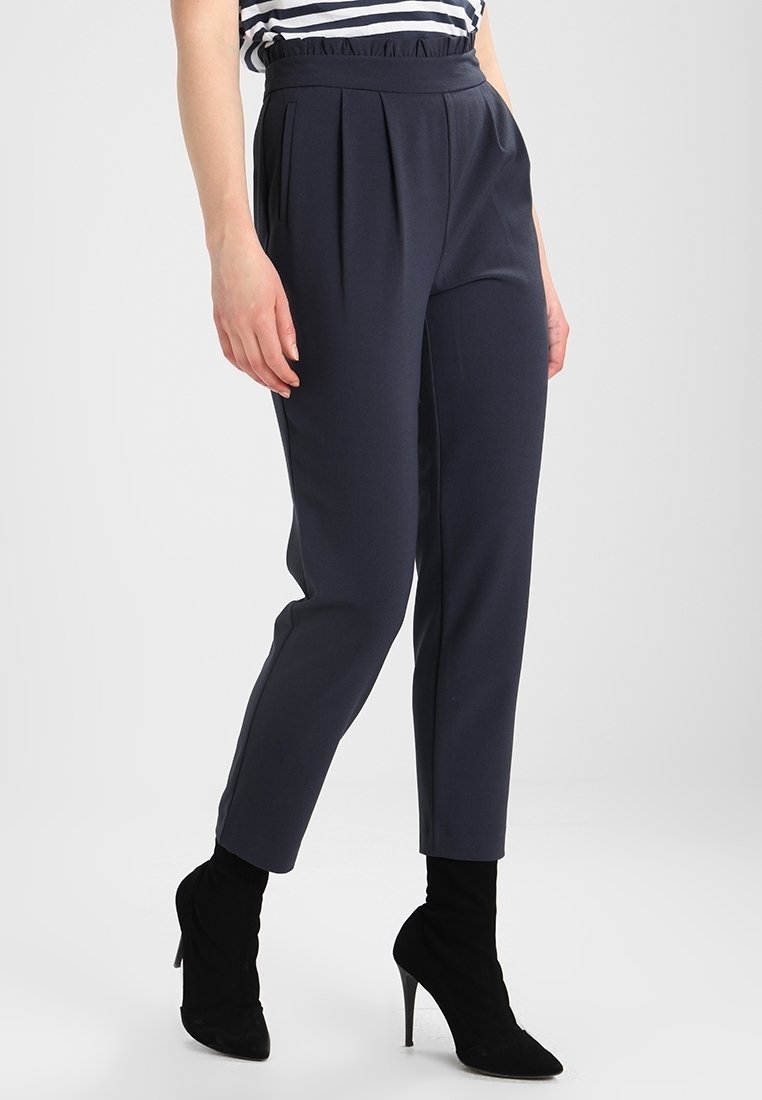 Femme portant un pantalon plissé bleu marine foncé taille haute, associé à des bottines noires à talons et un haut rayé, debout avec une jambe légèrement avancée.
