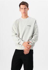 Sweatshirt cinza claro com gola redonda e punhos canelados, apresentando um pequeno logótipo preto "PEGADOR" no peito. Usado com jeans pretos.