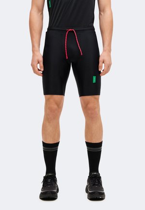 Homme portant un short de sport noir moulant avec un cordon rose, des chaussettes noires à rayures grises et des chaussures de course noires, debout les bras détendus.
