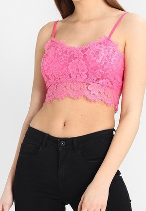 Kvinde iført en lyserød blomsterblonde crop top med tynde stropper og sort højersiddende jeans, torso og arme synlige mod en hvid baggrund.