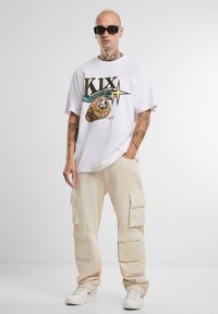 Hvit grafisk t-skjorte med fet "KIX" design og ringillustrasjon, kombinert med beige cargo-bukser med flere lommer og hvite sneakers.