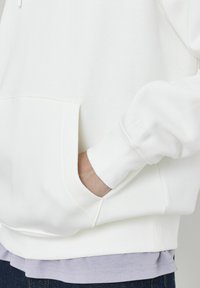 Sweatshirt en coton blanc avec une poche kangourou sur le devant, des poignets côtelés et une couche de chemise lavande à l'ourlet, avec une texture douce.