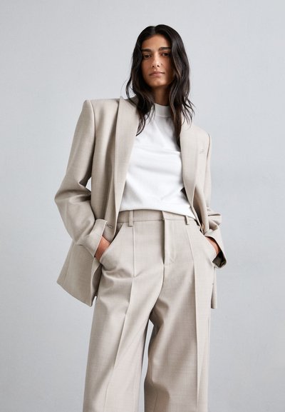 Beige, fazonra szabott öltöny, oversized blézerrel és magas derekú, pliszírozott nadrággal. Az anyag simának tűnik, finom textúrával; egy fehér felsővel párosítva.