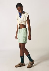 Crop top color crema con nodo frontale, pantaloni corti verde menta con tasche laterali, calze verdi e sneakers marroni con accenti neri.