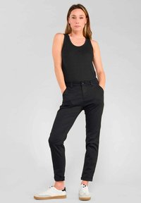 Zwarte tanktop en slim-fit zwarte broek van lichtgewicht stof. Model staat met handen in de zakken en draagt witte schoenen met tan-kleurige zolen.