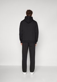 Sweatshirt à capuche noire et pantalon assorti dans une coupe décontractée. Le matériau semble doux avec une texture lisse. Des baskets blanches complètent le look.