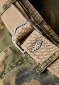 Cinturón de lona color canela con hebilla de metal plateado, unido a una tela de camuflaje verde, con detalles de costura y una pequeña etiqueta de logo.