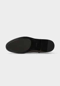 Semelle d'une chaussure de ville noire avec une bande en caoutchouc texturé au niveau du talon et des orteils sur un fond clair uni.