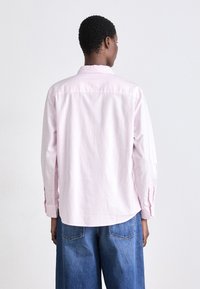 Camicia a righe rosa chiaro con maniche lunghe e polsini abbottonati, caratterizzata da un orlo curvo e un dettaglio sullo yoke posteriore. Abbinata a jeans a gamba larga blu.