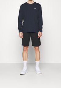 Marineblaue Langarmshirt mit Logo auf der Brust, kombiniert mit schwarzen Shorts. Das Model trägt weiße Sneaker und Socken. Einfaches und körpernahes Design.