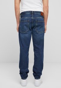 Jeans de mezclilla azul con un corte relajado, costuras visibles y dos bolsillos traseros. Llevados con zapatillas blancas, mostrando puños elásticos en los tobillos.