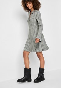 Vestido de punto gris con cuello, que cuenta con mangas largas y un dobladillo acampanado. Combinado con botas negras hasta el tobillo con hebillas y suelas texturizadas.