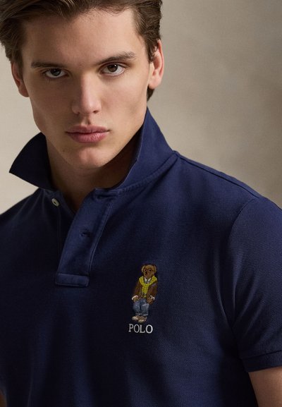 Polo bleu marine en coton texturé, avec un ours brodé portant une veste et un jean, accompagné du texte "POLO" en dessous du motif.