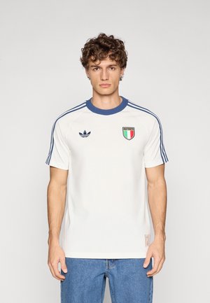 Camicia bianca a maniche corte con colletto blu e tre strisce blu sulle spalle, con una toppa della bandiera italiana e il logo Adidas sul petto.