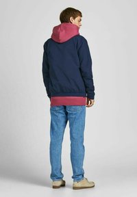 Marineblauwe jas met een roze capuchon, gelaagd over een bordeauxrode hoodie. Gecombineerd met blauwe jeans en lichtbeige schoenen. Glad materiaal, casual ontwerp.