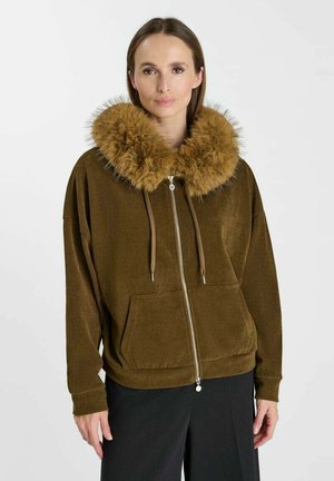 Donna con giacca marrone con cerniera, ampio collo in pelliccia soffice e coulisse, abbinata a pantaloni neri, su sfondo semplice.