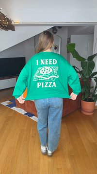 Άτομο με μακριά μαλλιά που φοράει πράσινο φούτερ με την επιγραφή "ΧΡΕΙΑΖΟΜΑΙ ΠΙΤΣΑ" και γραφικό πίτσας, στέκεται σε ένα σαλόνι με φυτά και έπιπλα.