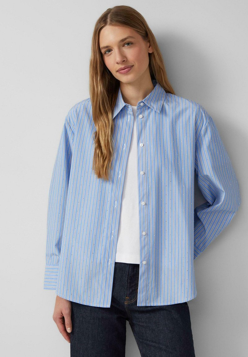 Camicia button-up blu chiaro con strisce verticali bianche e gialle, maniche lunghe e colletto classico. Indossata sopra un top bianco.