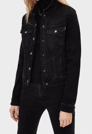 Kvinde iført en sort denimjakke med sølvknapper over en sort rullekrave og matchende sorte denimjeans.