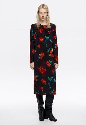 Mujer con un vestido midi negro con patrones florales en rojo y verde y botas negras hasta la rodilla, posando frente a un fondo blanco liso.