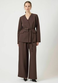 Bruine overslagjas met ceintuur in de taille en voorzakken, gecombineerd met wijde broekspijpen. Soepele stof, gestructureerd ontwerp en subtiele glans. Zwarte hakken.