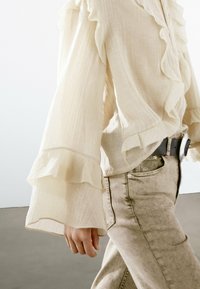 Light cream blouse with ruffled sleeves, textured fabric, and a relaxed fit, paired with light beige jeans and a black belt.
