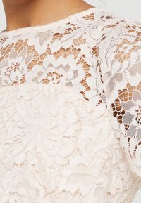 Detailní záběr bílé květinové krajkové blůzy, která ukazuje složité vzory a průsvitnou tkaninu na rameni a horní části hrudníku.