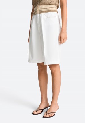 Persona con gambe snelle che indossa pantaloncini bianchi fino al ginocchio, top beige in maglia e sandali ciabatta neri con tacco, in piedi davanti a uno sfondo semplice.