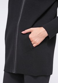 Veste noire côtelée avec fermeture éclair, poche latérale et tissu texturé. Coupe ajustée avec manches longues et ourlet droit.