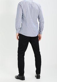 Modrobíle pruhované tričko s dlouhým rukávem a knoflíkovými manžetami, kombinované s černými úzkými kalhotami a černými botami, pohled zezadu.