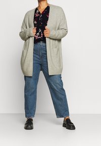 Person som bär en grå öppen cardigan, mörkblommig blus, blå jeans och svarta loafers, stående mot en neutral bakgrund.