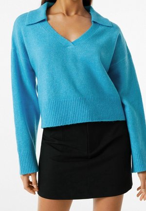 Donna che indossa un maglione azzurro chiaro a scollo a V con colletto e una minigonna nera su sfondo bianco.