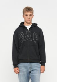 Ung man i en svart GAP zip-up hoodie med framfickor och ljusblå jeans, stående framför en helt vit bakgrund.