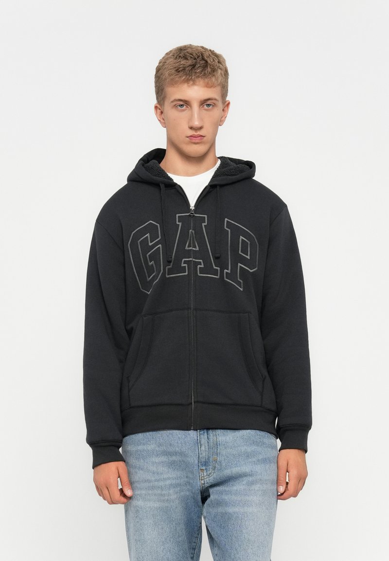 Ung man i en svart GAP zip-up hoodie med framfickor och ljusblå jeans, stående framför en helt vit bakgrund.