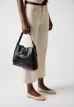 Personne portant un pantalon beige court et des ballerines crème tenant un sac à bandoulière en cuir noir brillant avec une boucle argentée et un petit logo.