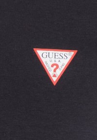 Tessuto nero con un logo triangolare rosso contenente il testo "GUESS®", "U.S.A." e "WASHED JEANS", messo in risalto da un punto interrogativo centrale.