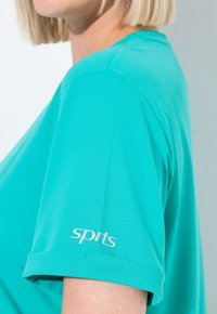 Detailní záběr osoby v tyrkysovém sportovním tričku s krátkými rukávy, s texturovanou tkaninou a logem "sports" na rukávu.