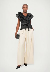 Zwarte blouse met ruches en een ceintuur in de taille, gecombineerd met beige broek met wijde pijpen. Het model houdt een kleine zwarte handtas vast en draagt zwarte hakken.