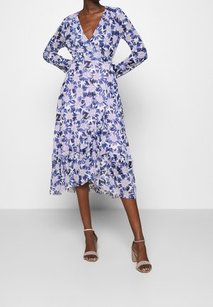 Frau trägt ein langärmliges, V-Ausschnitt, florales Wickelkleid mit lila und weißen Blumen, kombiniert mit beigen Sandaletten mit hohen Absätzen.