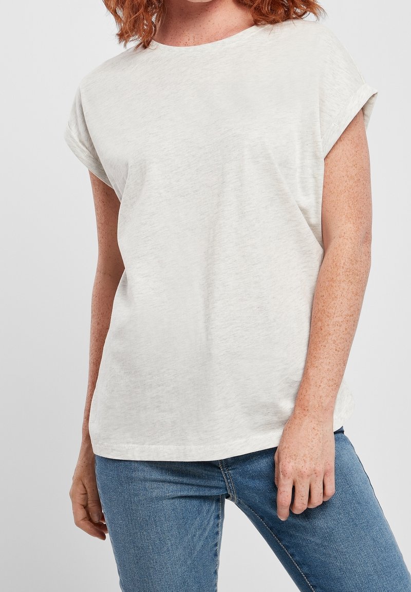 Femme à la peau claire avec des taches de rousseur, portant un t-shirt blanc uni à manches courtes et un jean bleu, debout devant un fond clair.