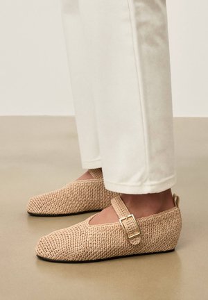 Bézs színű szövött Mary Jane cipők arany csattal, fehér nadrágot viselő lábakon, sima bézs színű padlón állva.