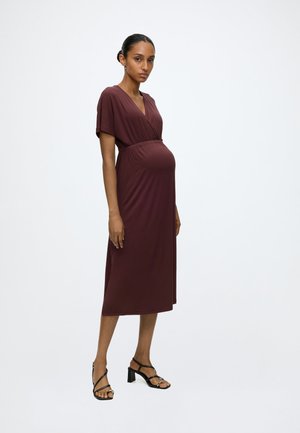 Femme enceinte portant une robe portefeuille marron longueur genoux et des sandales noires à talons, debout devant un fond blanc uni.